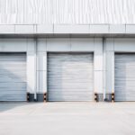 Modern industrial warehouse building exterior with three closed roller shutter doors. Grey facade and empty concrete loading dock area for logistics and shipping. Insulated lath roller shutters in level access openings